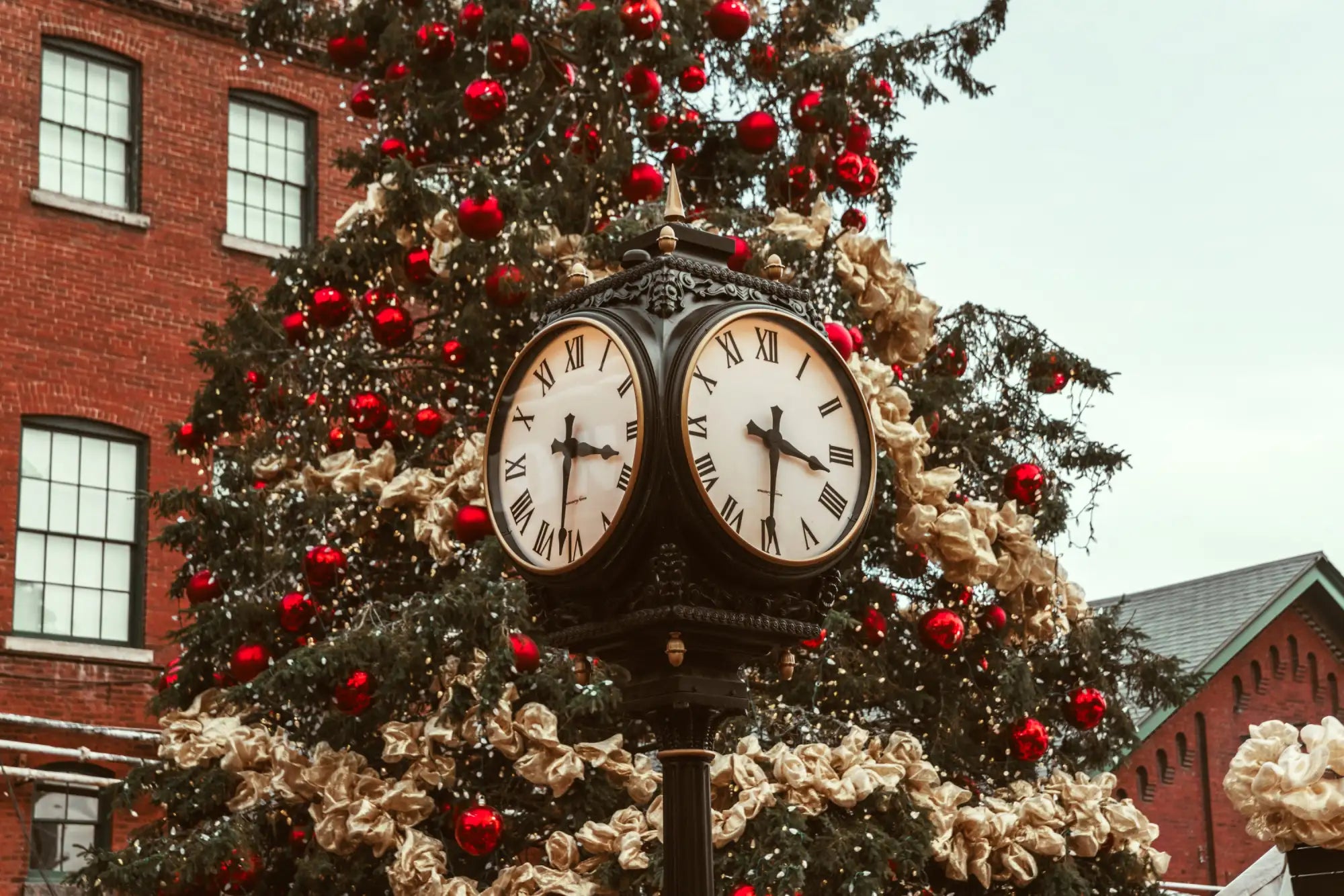 A black ornate street clock with two faces featuring white dials and roman numerals, mounted atop a decorative pole beside a christmas tree adorned with red ornaments and cream-colored floral garlands.