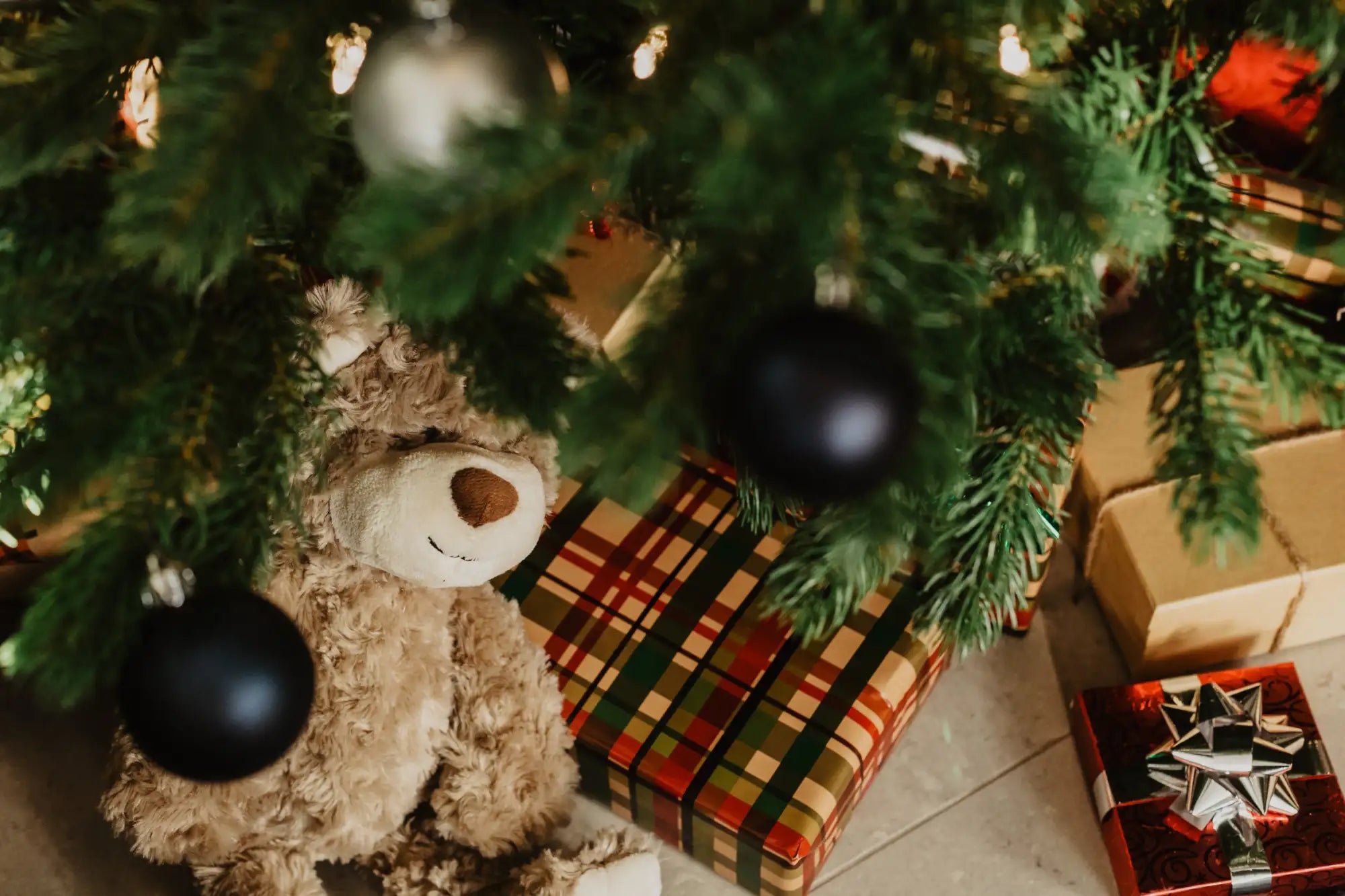 A soft, light brown plush teddy bear with a stitched smile and nose sits nestled under a christmas tree.